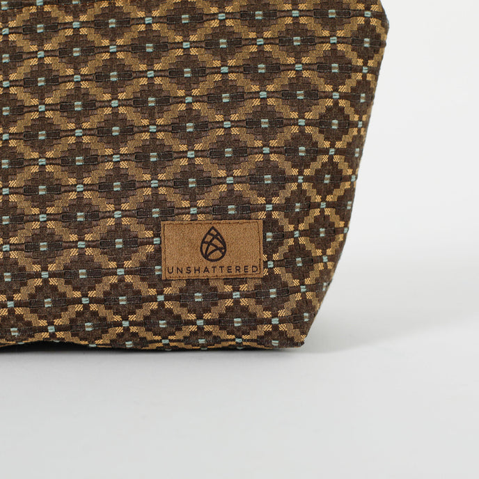 Close up of a brown, ultrasuede Unshattered label on a makeup bag with a brown and blue geometric pattern