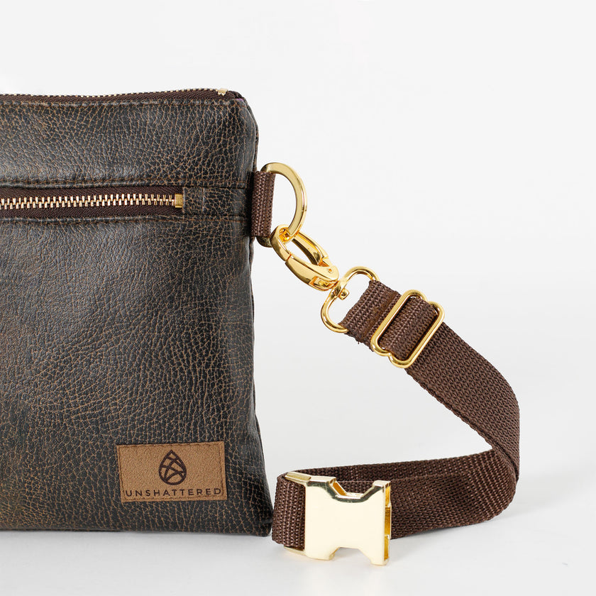 Close up of gold belt buckle on a brown belt strap attached to brown 3-in-1 bag on a white background