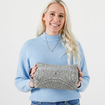 A woman wearing a blue sweater holding a gray spotted Haley toiletry kit with a geometric pattern, featuring a zip closure and a gray fabric lining.