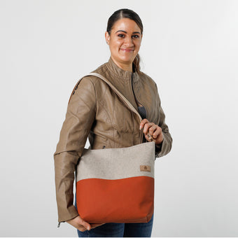 A woman wearing a  faux  leather brown jacket with a beige and orange tote bag with tan straps and gold hardware named after Deanna