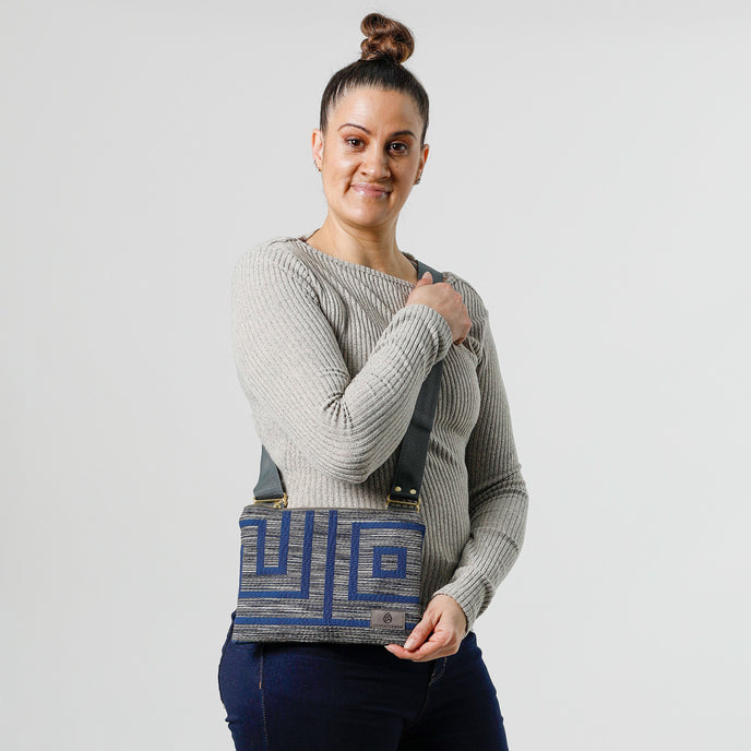 A woman carrying a blue and gray geometric patterned crossbody pouch with an adjustable strap and gold-tone hardware, displayed against a white background named after Lisa.