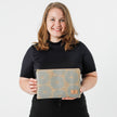A woman holding a large zip pouch with a warm tan base and vibrant blue flower pattern, featuring a gold-tone zipper  named after Sherry on the bottom right.