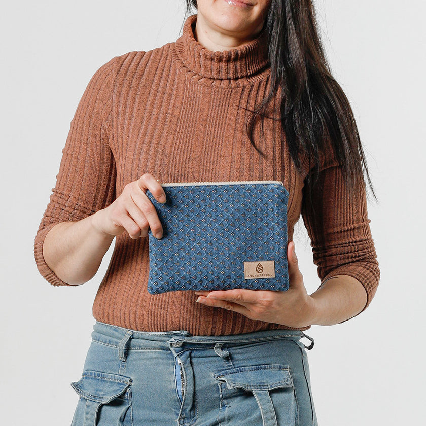 A woman holding a small zip pouch with blue paisley fabric and gold-tone hardware, featuring a brand tag that reads Unshattered named after Shiera