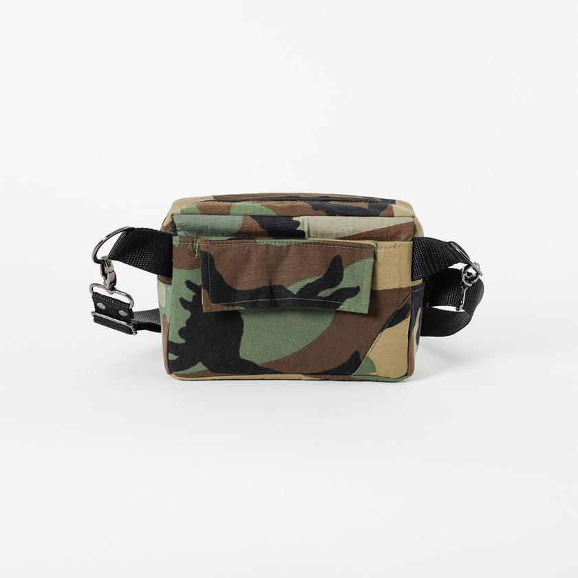 A pocket on the back of a woodland camo errand bag with black straps and black metal hardware on a white background 