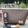 Light brown makeup bag with makeup on a stone wall in front of a flower garden