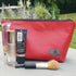 A red makeup bag by Unshattered with makeup brushes and concealer on a stone wall in front of a garden