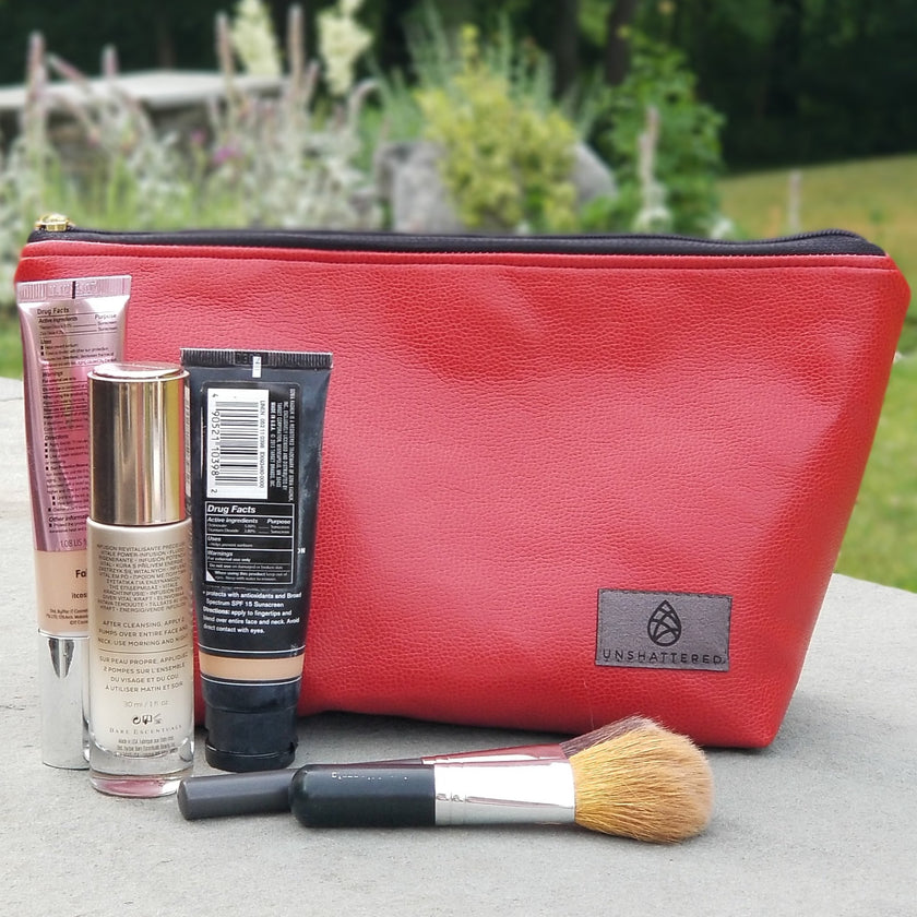 A red makeup bag by Unshattered with makeup brushes and concealer on a stone wall in front of a garden