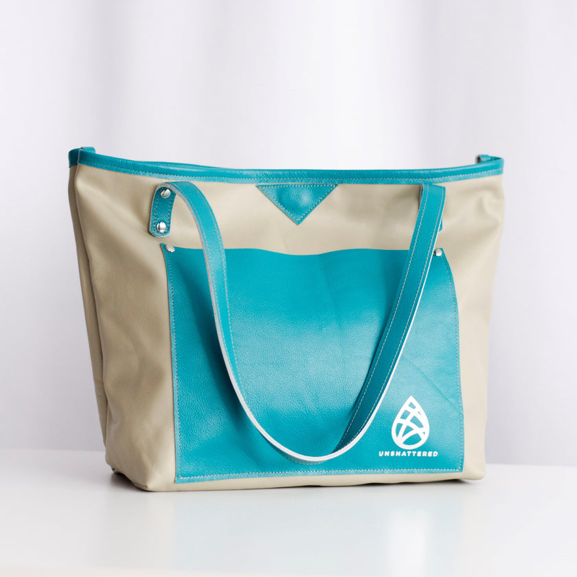 A leather tote bag with pebble-colored body and cerulean-colored trim, featuring two shoulder straps and a logo on the side.