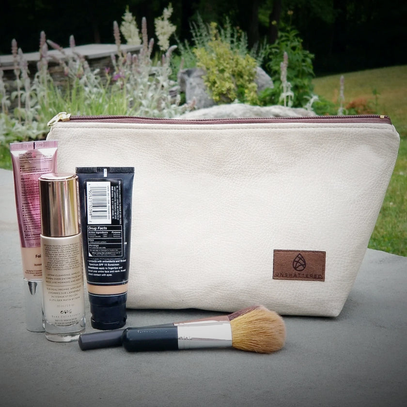 A white makeup kit with a zip closure, made from industrial luxury leather alternative with makeup on a stone wall in front of a flower garden